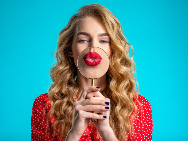 金发女郎拿着一个放大镜在她的红唇前。用放大镜放大一个大红唇的女孩。微笑。漂亮模特在调情。一个年轻女子拿着一个放大镜放在嘴唇前。图片下载