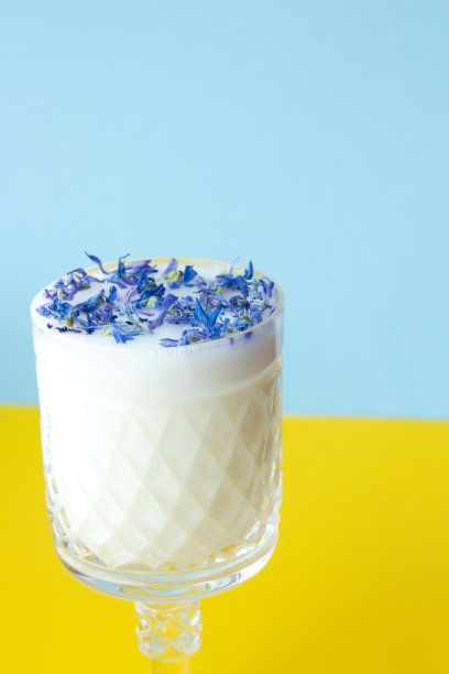 美丽的水晶玻璃与牛奶和蓝色花瓣在蓝色和黄色的背景。花在牛奶图片下载