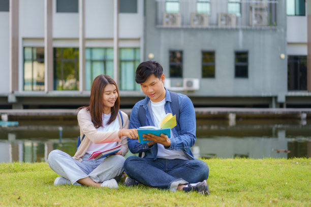 亚裔男女大学生坐在校园草地上看着镜头，微笑着，在公园里一起看书。图片下载