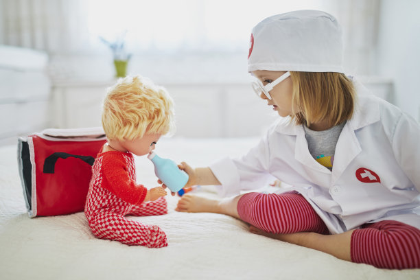 可爱的蹒跚学步的小女孩在白大褂扮演医生，给她的洋娃娃医疗护理图片下载