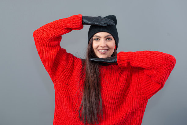 十几岁的女性戴着一顶黑色的冬季帽子和皮手套，穿着一件红色毛衣，孤立在灰色图片下载
