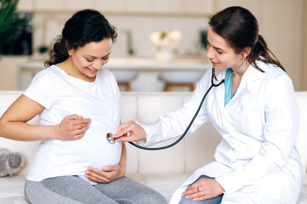 体贴的医生检查婴儿的心跳。快乐的混血孕妇坐在沙发上，接受医生检查，在家或医院，抚摸着自己的肚子。健康怀孕的概念图片下载