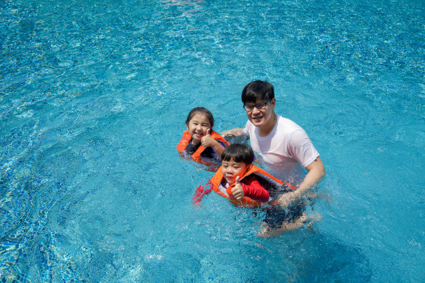 一位亚洲父亲教他的儿子和女儿在游泳池里游泳。姐姐和弟弟穿着橙色的救生衣，微笑着竖起大拇指。图片下载