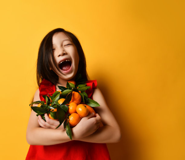 穿红衬衫的亚洲小孩。大声笑着，抱着橘子和橘子，在橙色的背景下摆姿势。近距离图片下载