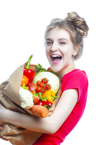 年轻的白人女性在红色衬衫拿着装满多种蔬菜和杂货的生态纸袋在白色背景下摆姿势。垂直的形象定位图片下载