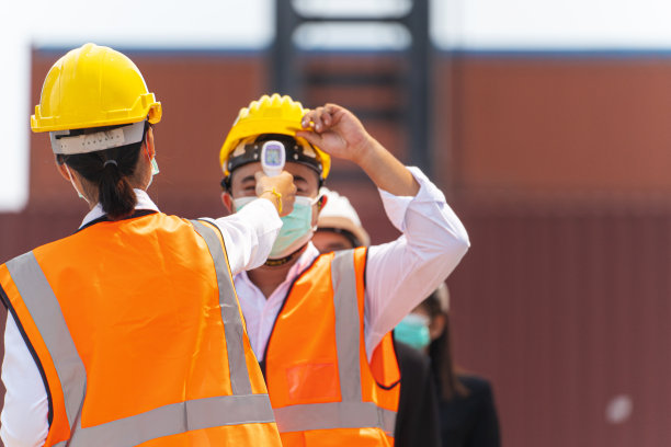 工厂女工戴着医用口罩，穿着安全服，用非接触式红外温度计测量排队工人的体温。货物集装箱的背景。图片下载