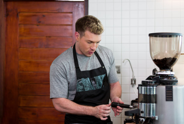 咖啡店的咖啡师穿着围裙，准备咖啡研磨机倒在咖啡店里，开心地微笑着。工人在蒸汽咖啡机煮咖啡图片下载