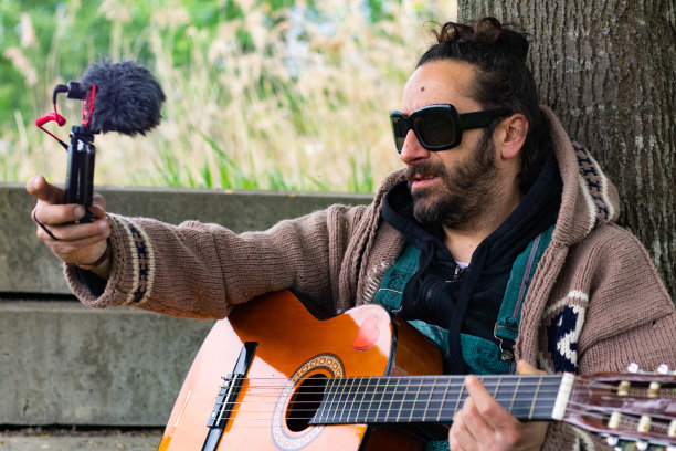 一个留着胡子戴着眼镜的嬉皮士自己在户外的草地上弹吉他拍视频图片下载