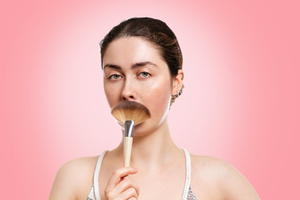 一个年轻美丽的女人的肖像，严肃地拿着毛绒绒的胡子形状的刷子。粉红色的背景。化妆、美容、美容的概念图片下载