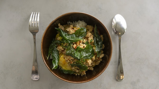 米饭配上炒好的猪肉和罗勒，放在一个棕色的碗里，勺子和叉子都是不锈钢的，灰色背景，俯视图图片下载