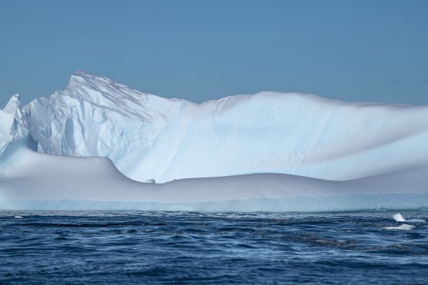 一个巨大的冰山，有光滑的，弯曲的边缘和一个粗糙的，崎岖的山峰，漂浮在深蓝色的水域下，蓝天图片下载