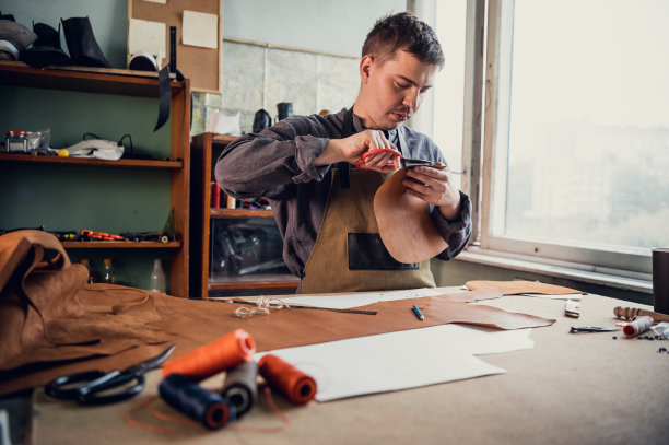 一个年轻的制革工人在桌子上用皮革做鞋坯，桌子上是线和剪刀图片下载
