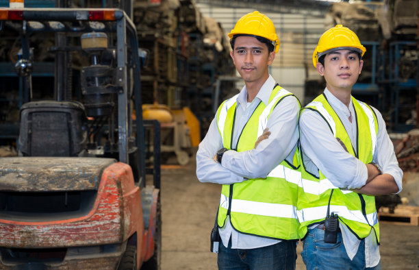 两名身穿制服、头戴头盔的年轻亚洲工程师站在那里，拥抱着胸膛，看着镜头，看着工业商业概念中的汽车配套件工厂。图片下载