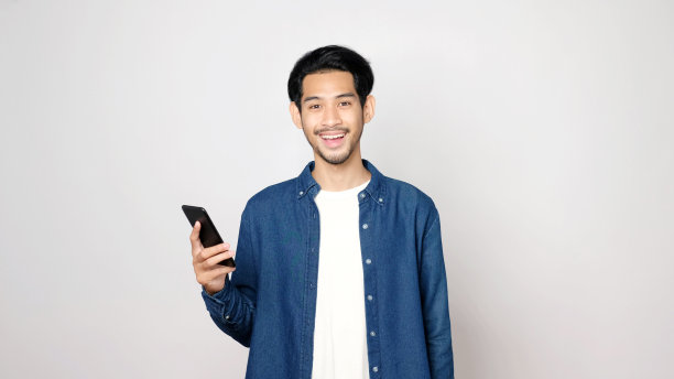 亚洲人拿着手机微笑着看着相机，孤立地站在灰色背景上，智能手机上的人，科技生活方式，电子商务，在线商务，e学习，教育，物联网的概念图片下载