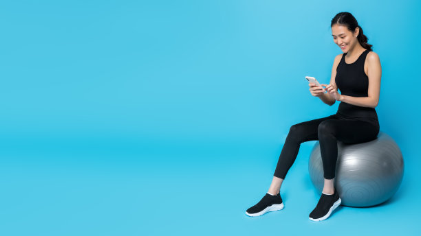 亚洲美丽快乐的女人拿着智能手机，坐在健身球后，孤立在蓝色背景白色工作室拍摄。概念苗条和健康的女孩锻炼。图片下载