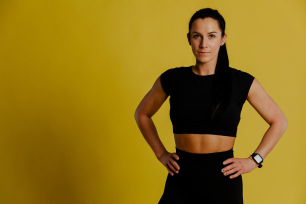 美丽的年轻健康的女人在黄色的背景，穿着黑色运动服，看着相机。图片下载
