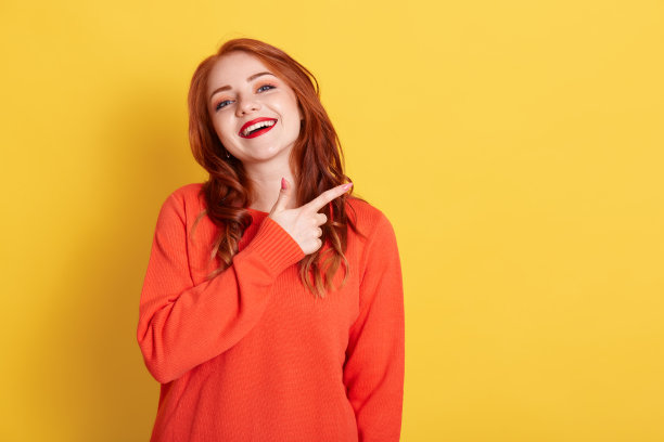 年轻女子的肖像与红头发指向食指一边，显示复制空间的推广，穿着休闲毛衣，看着相机与快乐的面部表情，孤立在黄色的墙。图片下载