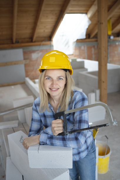 年轻漂亮的女砖匠戴着黄色的安全帽，正在房子里的建筑工地上锯砖图片下载