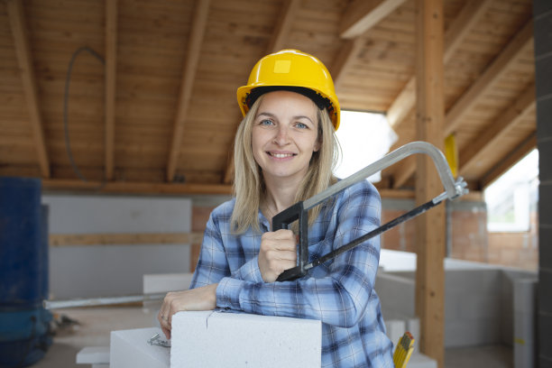 年轻漂亮的女砖匠戴着黄色的安全帽，正在房子里的建筑工地上锯砖图片下载