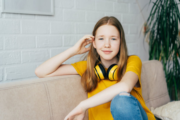 一个红发蓝眼少女的肖像，她坐在家里的沙发上，戴着无线耳机。图片下载