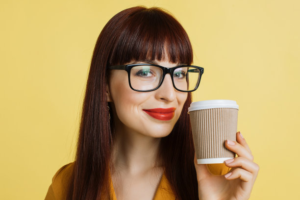 人、情感和热饮。近距离的可爱微笑迷人的白人女商人戴眼镜和亮红色的口红摆姿势与咖啡的黄色背景图片下载