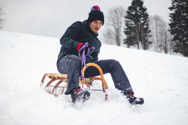 一名身着黑色冬装的成年男子坐在木制雪橇上高速行驶，并努力不从雪橇上摔下来。脚制动。用新鲜的粉末快速骑行。捷克共和国的冬季图片下载