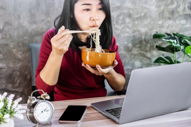 忙碌的亚洲女人一边吃面条一边在办公桌上用笔记本电脑工作图片下载