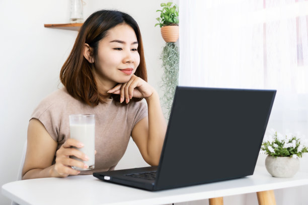 一位健康的亚洲女性在办公室边喝牛奶边用手提电脑工作，手里拿着一杯牛奶图片下载