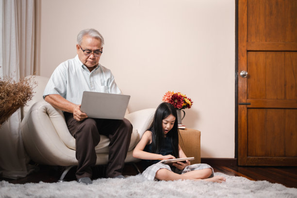 亚洲退休祖父使用笔记本电脑，坐在沙发上，而漂亮的孙女在客厅用平板电脑做作业。家庭教育家庭观念。技术和教育。图片下载