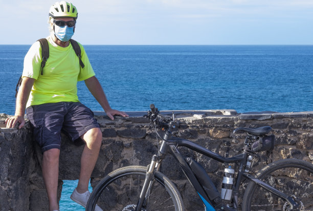 冠状病毒一名白发老人因冠状病毒感染戴着防护面罩，骑着自行车，戴着头盔，在户外活动，享受大自然和大海图片下载