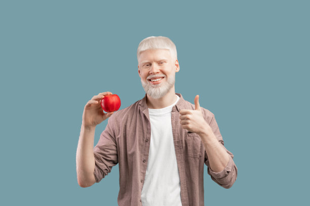 健康的生活方式。快乐的年轻白化病男子拿着红苹果，手势拇指向上和微笑，绿松石工作室的背景图片下载