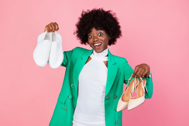 非洲女性在运动鞋和高跟鞋之间选择鞋子，粉色背景图片下载