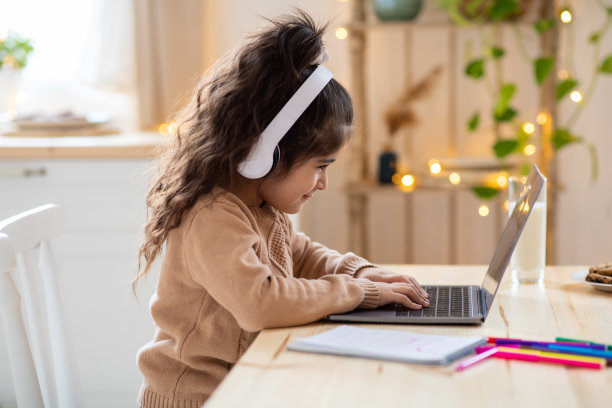 在线课程：在厨房里戴着耳机使用笔记本电脑的可爱小女孩图片下载