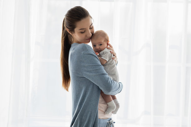 美丽的年轻母亲抱着她可爱的新生婴儿站在窗前图片下载