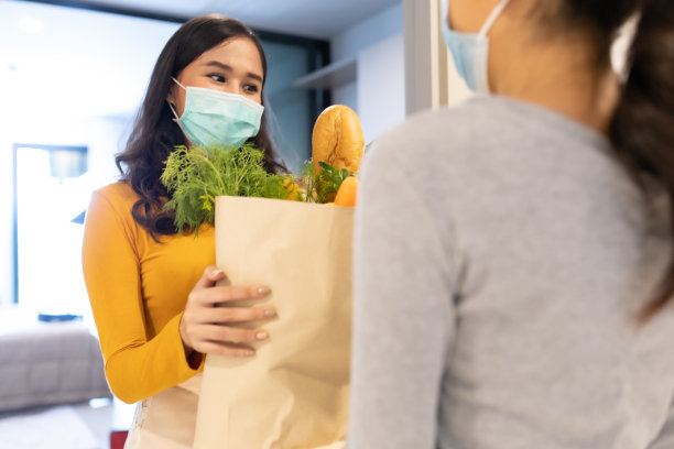 亚洲送人戴着口罩在红色制服处理袋食品，水果，蔬菜给公寓女顾客。新冠肺炎期间的邮差杂货送货服务。预防病毒感染的新正常生活方式概念。图片下载