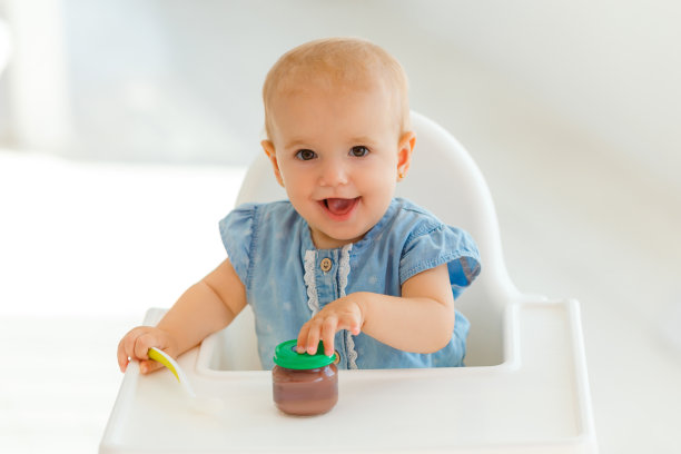 非常可爱和有趣的宝宝用勺子吃孩子的营养-水果泥。微笑的小女孩坐在白色的喂食桌上。第一有机儿童食品和家庭护理理念图片下载