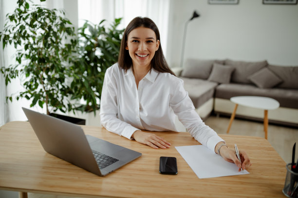 照片上的开朗漂亮的女学生微笑着用笔记本电脑在客厅做作业。图片下载
