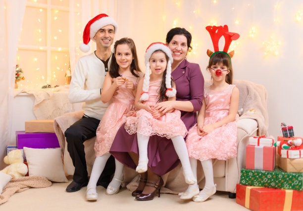 一个家长和孩子的全家福，穿着圣诞助手帽，坐在沙发上，在家里装饰着圣诞灯和新年节日礼物图片下载