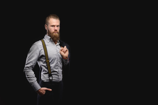 这是一位蓄着胡须的白人男子的肖像，他穿着时髦的西装和衬衫，肩上扛着夹克，看着镜头。在黑色的背景上。图片下载