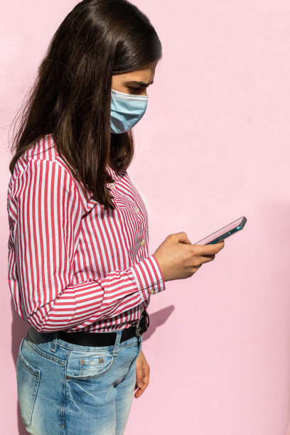 长头发的少女面罩covid - 19条纹衬衫牛仔裤使用手机上网发信息，聊天和浏览淡粉色背景和选择性聚焦的照片图片下载