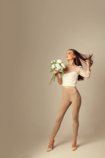 全身像的年轻白人褐发女人在露脐装和紧身裤与鲜花摆姿势和看着相机在米色的背景图片下载