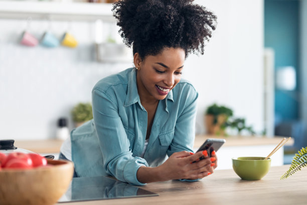 美丽的非洲裔女子一边用手机发短信，一边用筷子在厨房里吃面条。图片下载