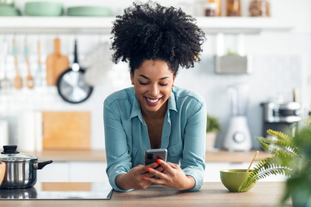 美丽的非洲裔女子一边用手机发短信，一边用筷子在厨房里吃面条。图片下载