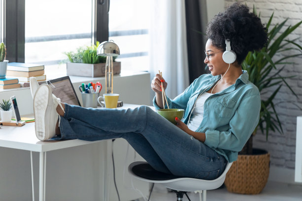 年轻漂亮的非洲裔商务女性在家里的办公室里用笔记本电脑打视频电话，用筷子吃面条。图片下载