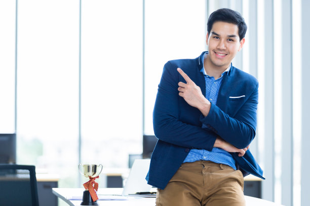 成功的感觉胜利者尖叫英俊年轻的亚洲商人拿着冠军杯，并显示拇指在办公室的背景。图片下载
