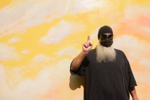 夏季肖像的成熟男子长灰胡子对云背景墙戴口罩，以防止Covid-19手指向上图片下载