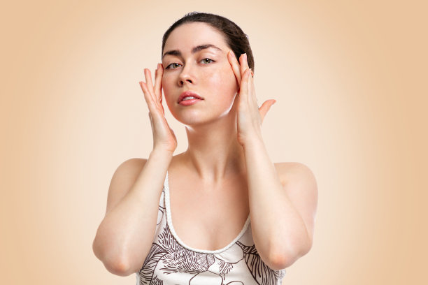 一个年轻女子的肖像与美丽的皮肤，收紧皮肤在她的太阳穴与她的双手。米色背景。副本的空间。美容、年轻化、美容的概念图片下载