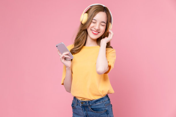 一名欢快的年轻亚洲女子戴着耳机听着音乐，享受着智能手机上最喜爱的播放列表应用程序，在粉红色的背景上跳舞。图片下载