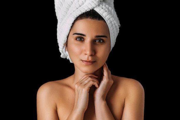漂亮的年轻女子，她的脸上有干净清新的皮肤。面对治疗。美容、美容和水疗。一个年轻美丽的女孩的肖像，头上戴着一条毛巾，在一个黑色的背景图片下载