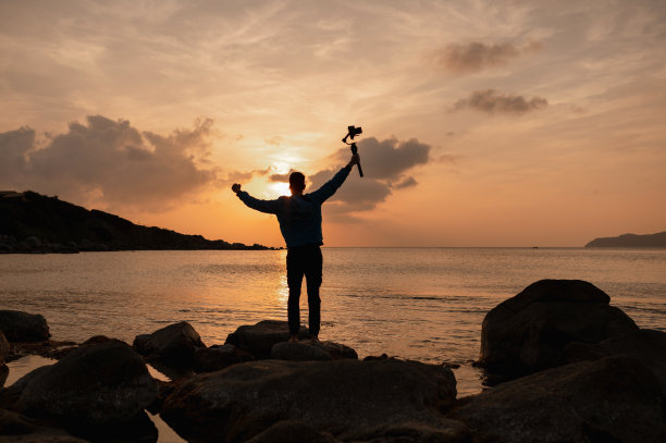 在海岸边的岩石上的人的剪影持有一个平衡环稳定器来拍摄日落的美丽风景。图片下载